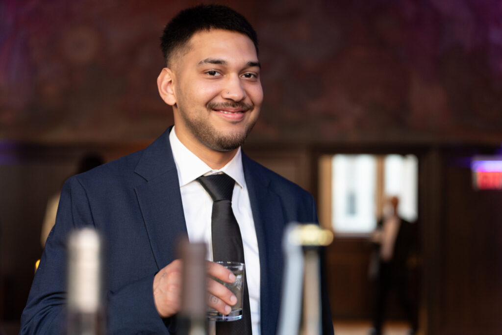 A man in a suit smiles while holding a glass, with blurred figures in the background.