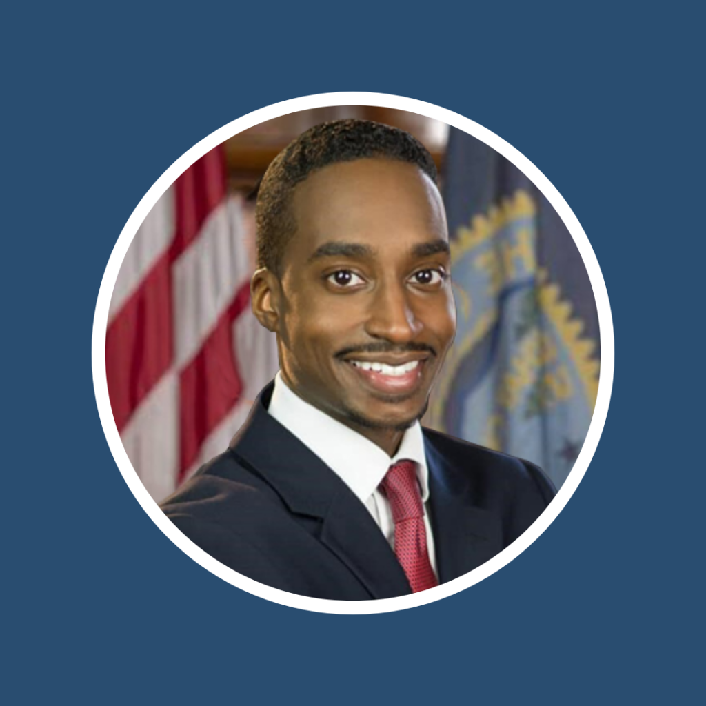A smiling man in a suit poses against a backdrop of flags and emblems.