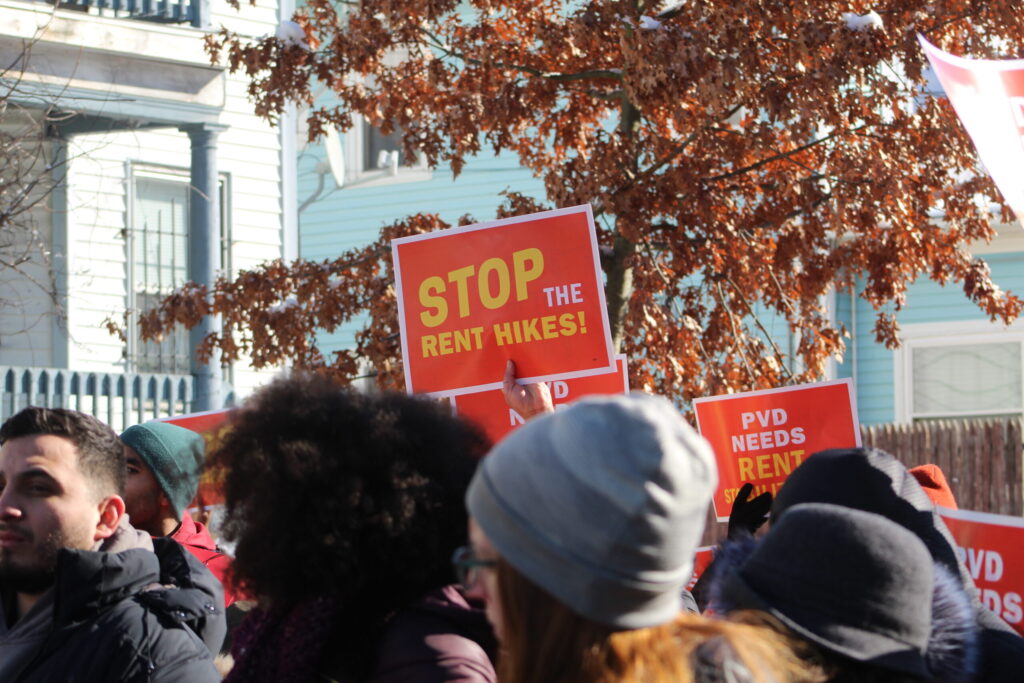 A crowd holds signs demanding to stop rent hikes, with trees and buildings in the background.