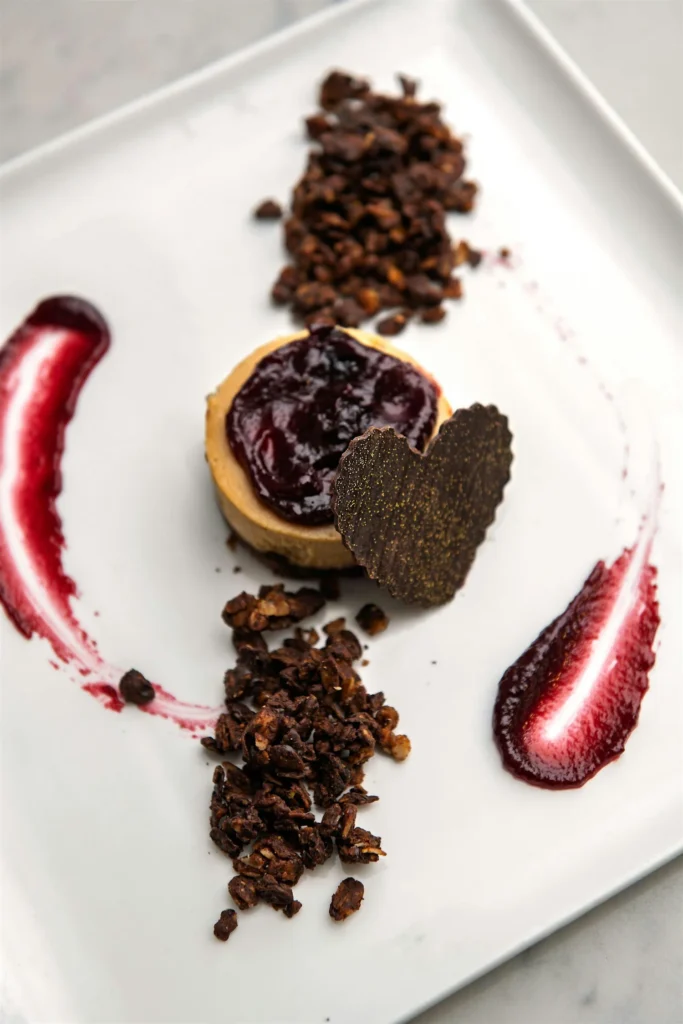 A dessert plate features a cheesecake topped with berry sauce, chocolate heart, and granola clusters.