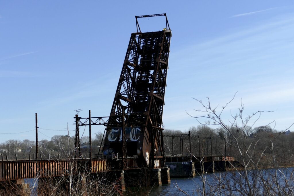 A rusted drawbridge stands at an angle over a river, surrounded by bare trees and a clear blue sky.