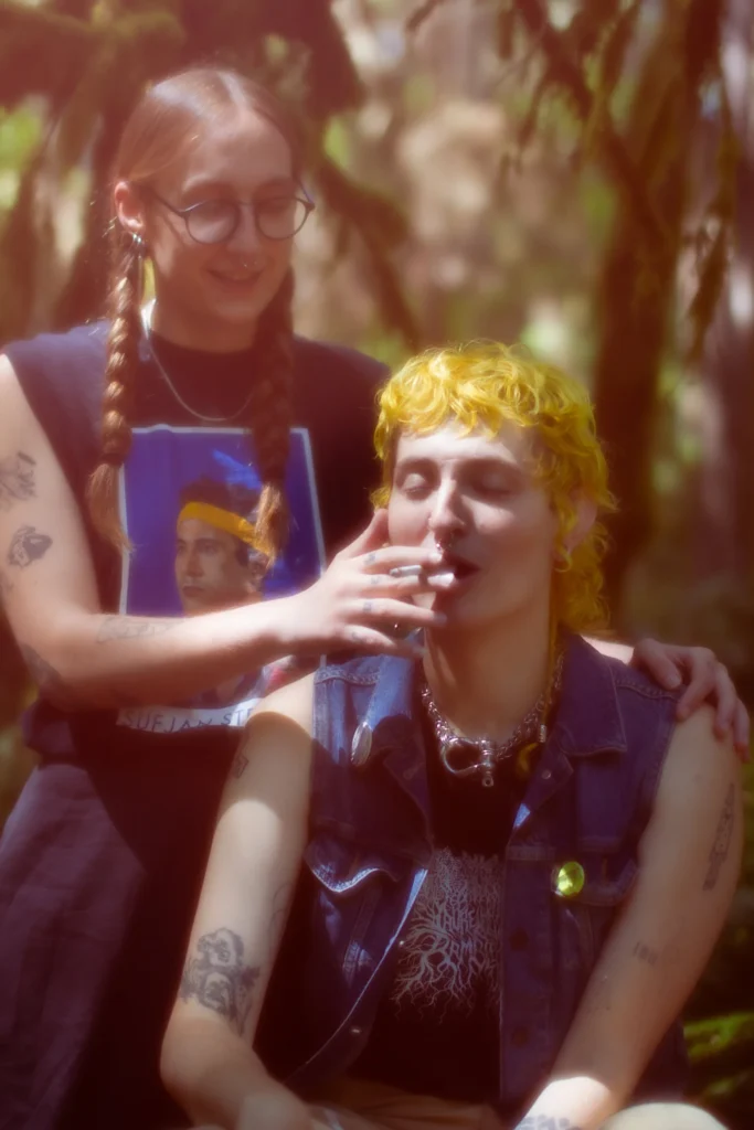 Two people interact in a forest. One smiles and touches the other's face, who is smoking. Both have visible tattoos.