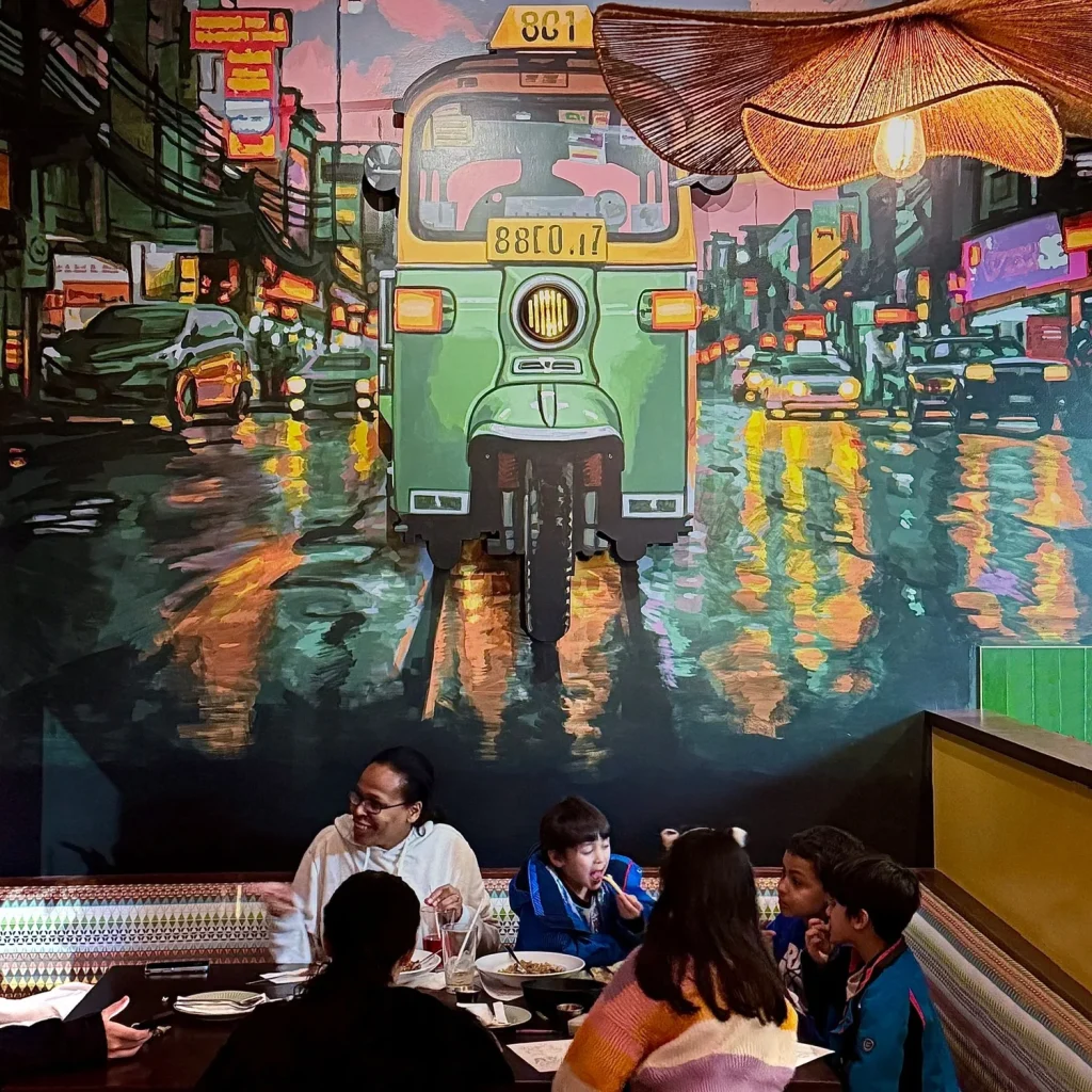 A group of people dining at a restaurant, with a colorful mural of a green tuk-tuk in the background.