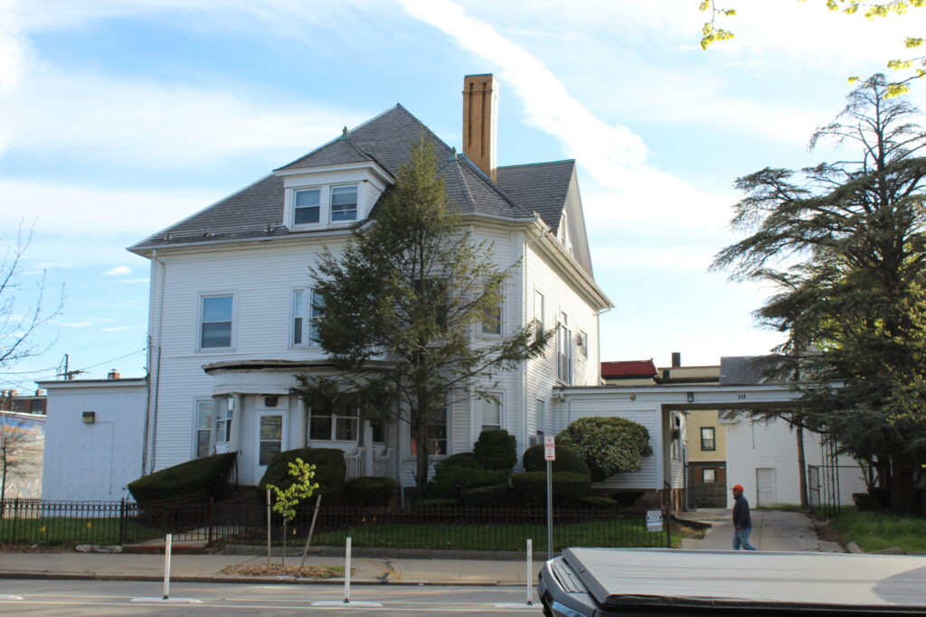 A large white house with a steep roof, surrounded by greenery, and a person walking nearby.