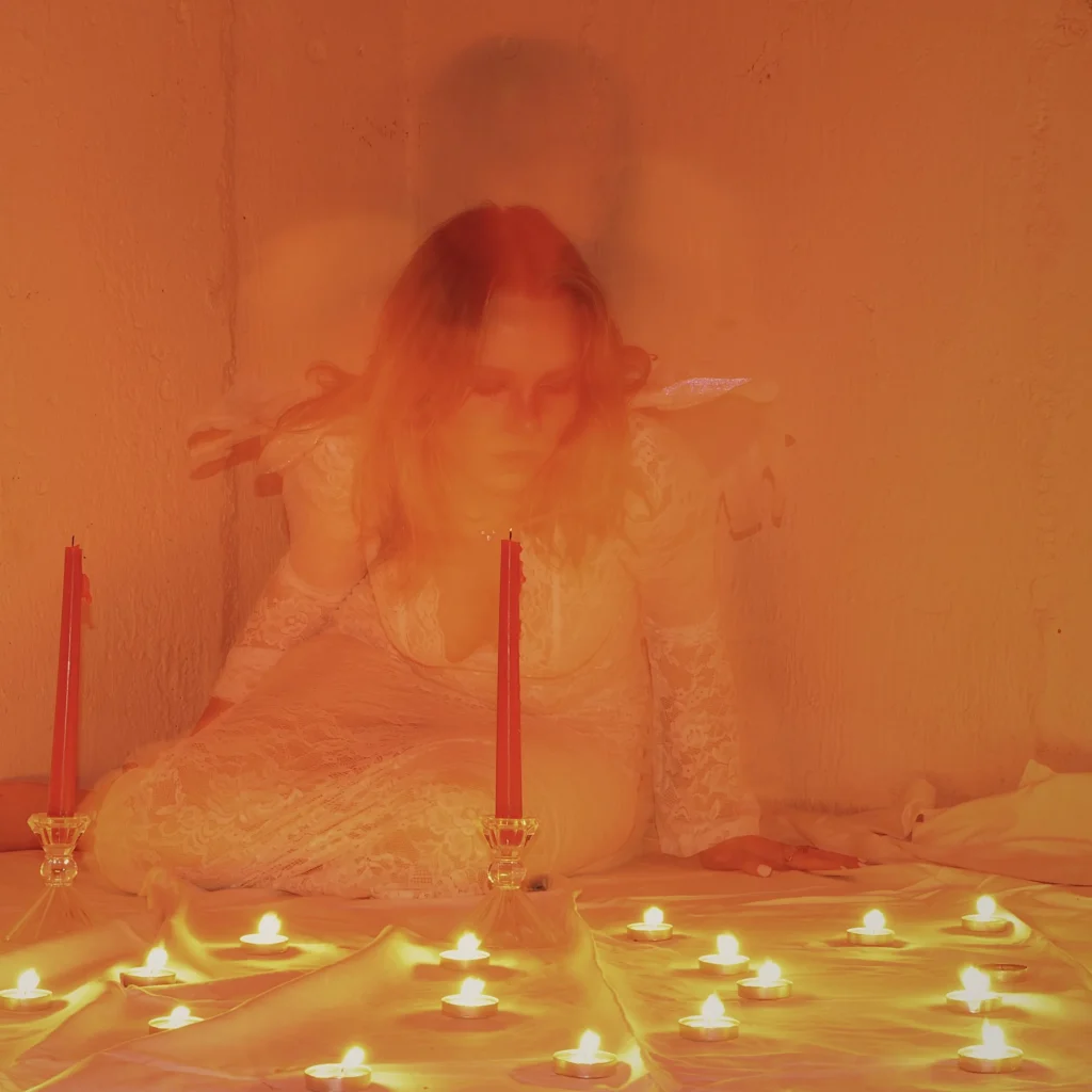 A woman in a white dress kneels among lit candles, surrounded by an orange glow.