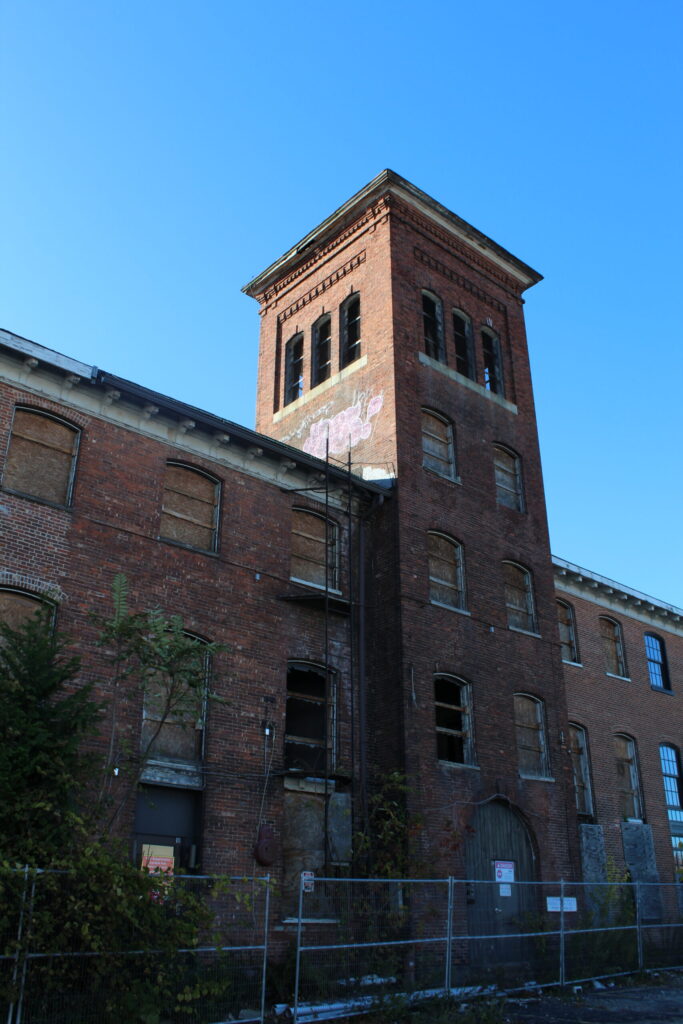 Abandoned brick building with boarded windows and a tall tower against a clear blue sky.