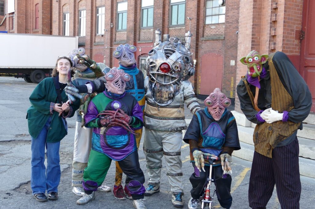 A group of six people in colorful alien costumes poses playfully outside a brick building.
