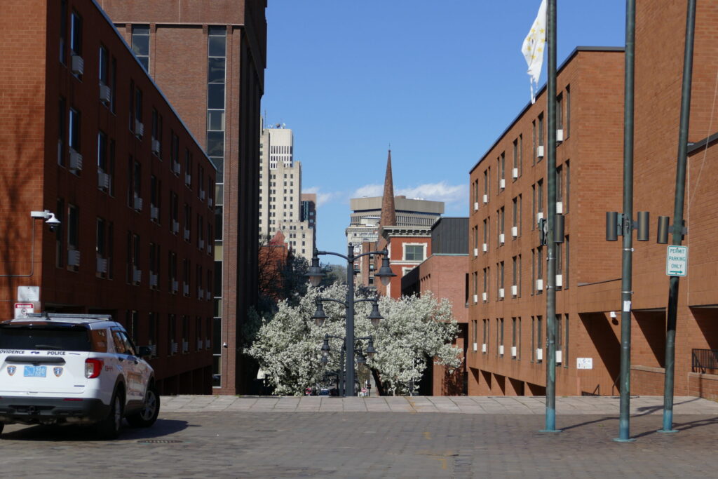 View of a city street lined with brick buildings, flowering trees, and a police vehicle parked nearby.