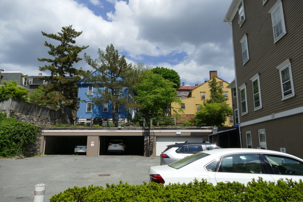 Blue and yellow houses behind a parking area with several cars and trees under a partly cloudy sky.