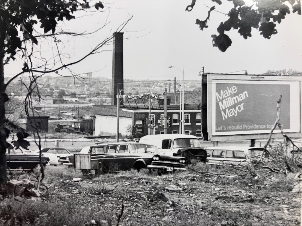 Overgrown lot with parked cars and a billboard promoting Millman for Mayor in Providence.