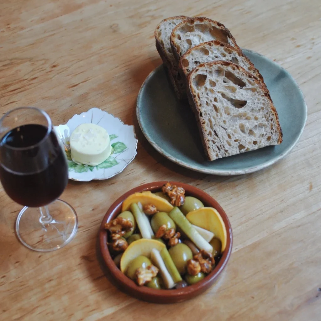 Sliced bread on a plate, a glass of dark liquid, butter, and a bowl of olives and snacks on a wooden table.