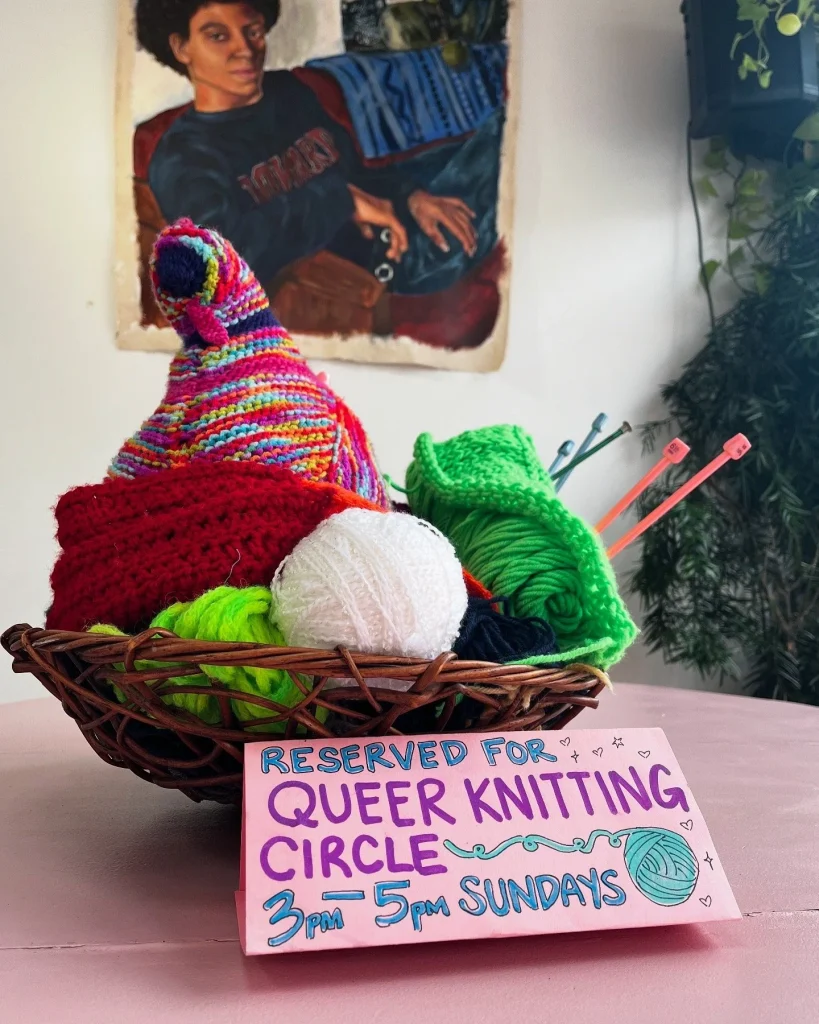 A basket of colorful yarn and knitting supplies with a sign for a Queer Knitting Circle.