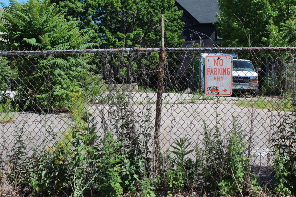 Chain-link fence with overgrown plants; a "No Parking" sign is visible, and a vehicle is parked behind it.