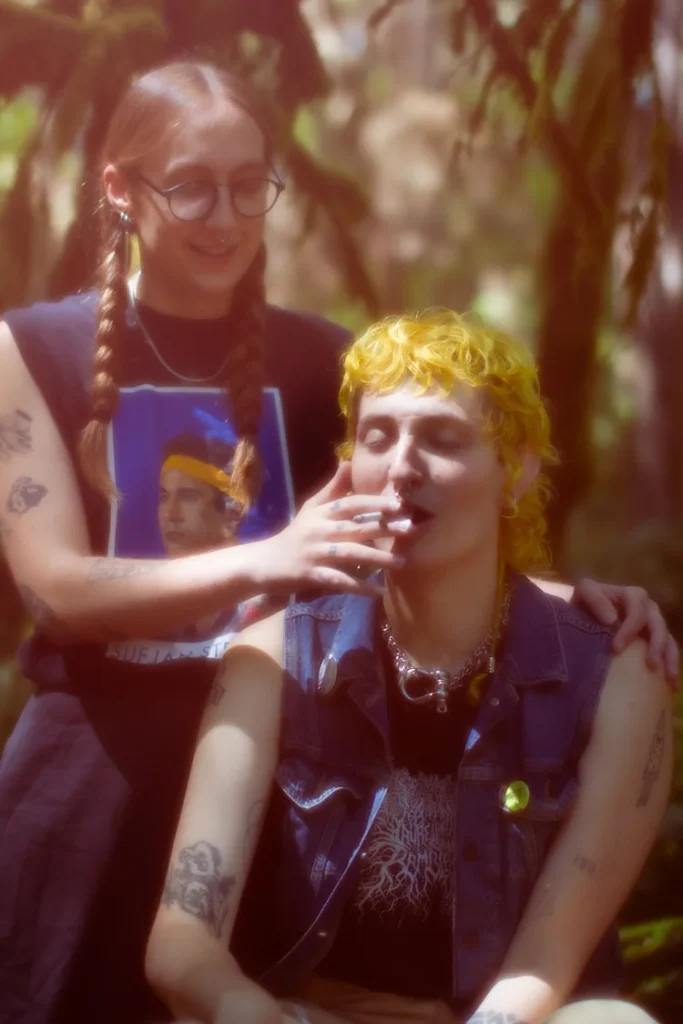 Two people in a forest; one offers a cigarette to the other, who is seated and smoking.