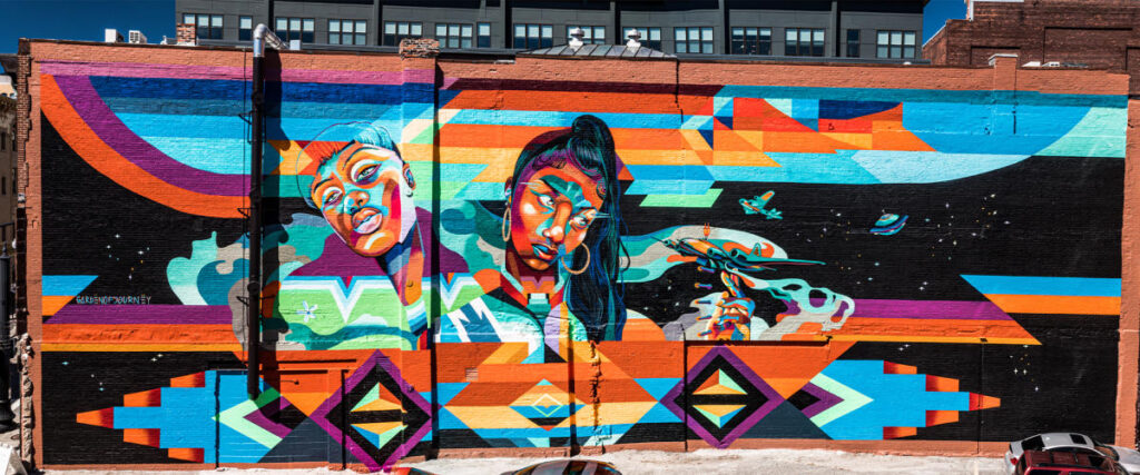 Vibrant mural featuring two women, colorful geometric patterns, and cosmic elements on a brick wall.