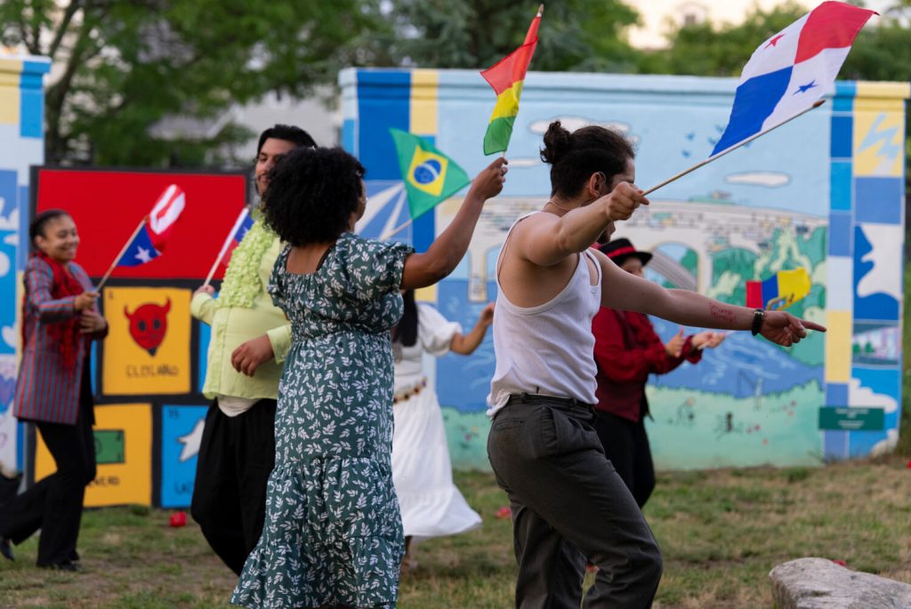 People dance in a grassy area, waving flags from various countries against a colorful mural backdrop.