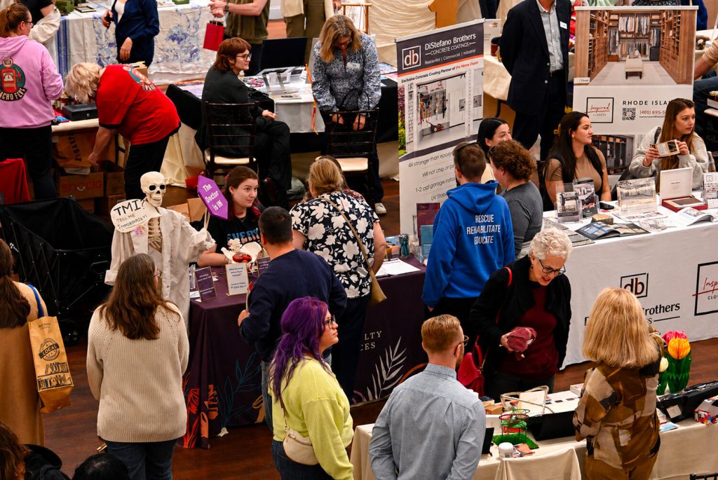Crowd at a market event, vendors display products, a skeleton figure holds a sign, people engage in conversation.