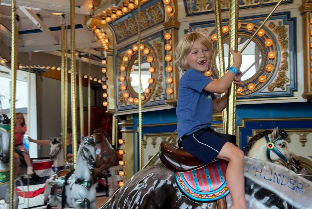 A child with blonde hair smiles while riding a carousel horse, surrounded by colorful lights and other riders.