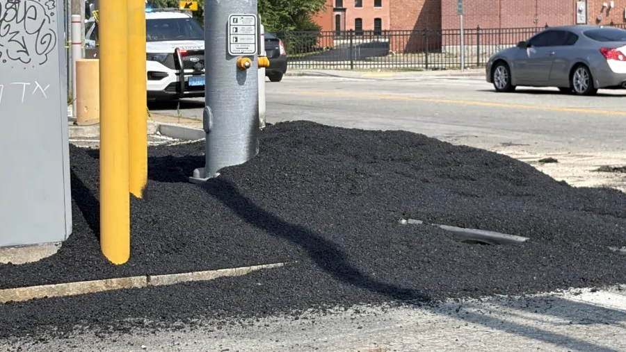 Asphalt pile near a traffic signal, with vehicles parked and a brick building in the background.