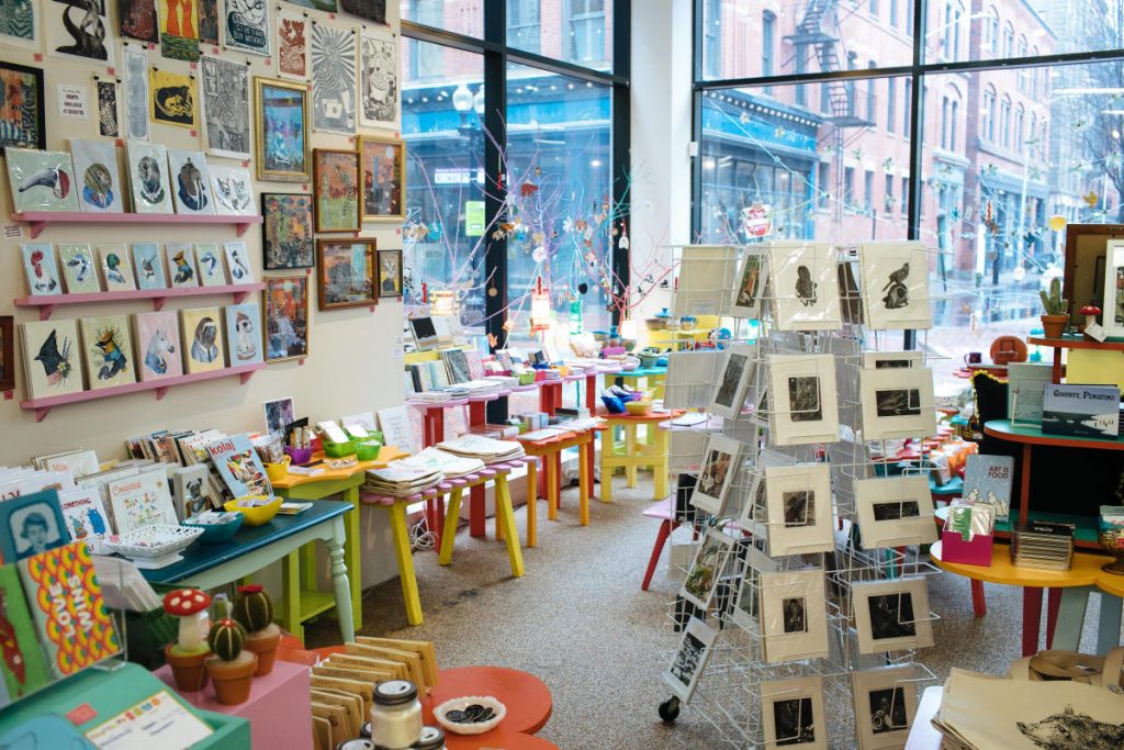 Colorful craft store interior filled with art prints, greeting cards, and decorative items on tables and walls.