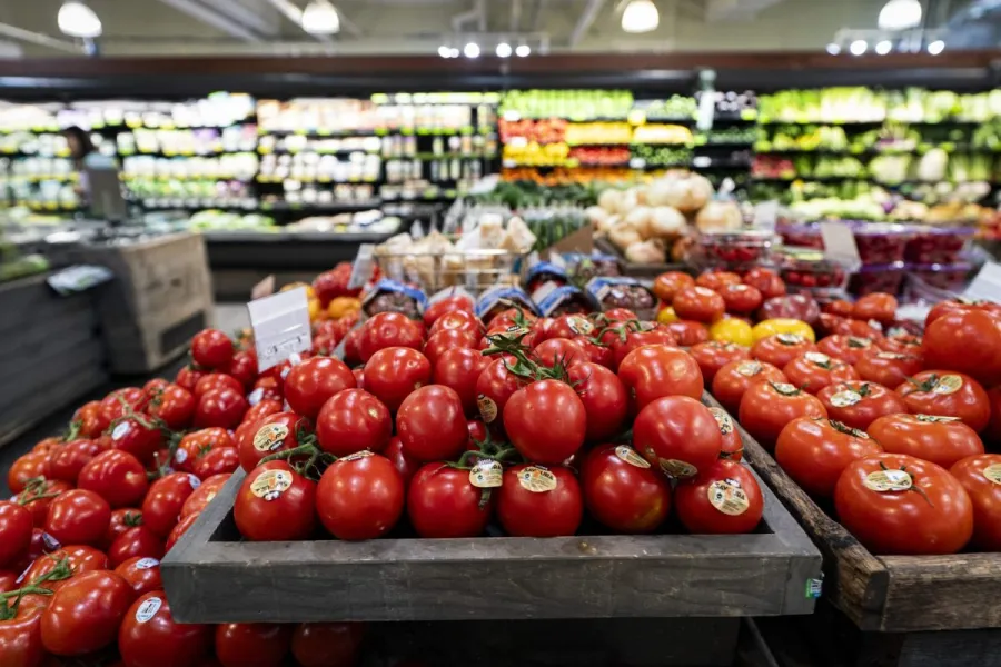 Red tomatoes are displayed in wooden crates, surrounded by various vegetables in a grocery store.